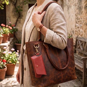 Könnte beinhalten: Eine braune Ledertasche mit einer kleinen roten Geldbörse. Die Tasche hat eine strukturierte Oberfläche und braune Griffe. Eine Person hält die Tasche und trägt einen beigen Blazer und eine braune Uhr.