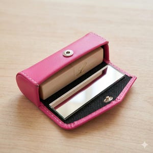 Könnte beinhalten: Ein leuchtend pinkfarbenes Lippenetui aus Leder mit einem silbernen Druckknopfverschluss, das einen Lippenstift und einen kleinen Spiegel enthält. Der Lippenstift hat ein goldfarbenes Etikett mit dem Buchstaben "L". Das Etui ist geöffnet und zeigt den Inhalt.