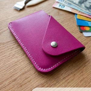 May include: A magenta hand-stitched leather cardholder with white stitching and a silver snap closure. The wallet is designed to hold six cards and cash. The image also shows a leather tool, a bone folder, and currency.