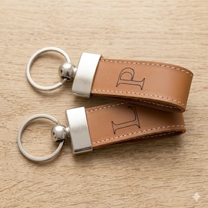 Puede incluir: Dos llaveros personalizados de cuero con herrajes plateados. Los llaveros son de color marrón claro con las letras "P" y "L" grabadas. Están sobre una superficie de madera marrón claro.