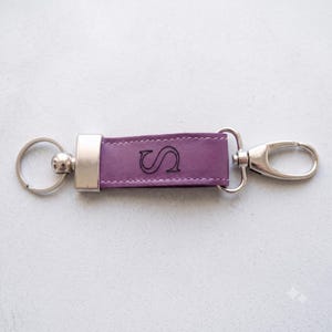 Puede incluir: Un llavero de cuero morado con una anilla de metal plateado en un extremo y un mosquetón plateado en el otro. La correa central de cuero morado tiene grabada la letra S en una fuente serif negra y cuenta con costuras de color blanco.