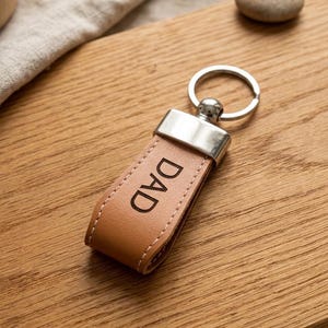 May include: A tan leather keychain with the word "DAD" printed in black. The keychain has a silver-coloured metal top and a silver key ring. The item is on a wooden surface.