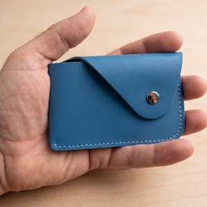 May include: A blue leather card holder with a flap closure and a silver snap. The edges are stitched with white thread. The card holder is held in a hand.
