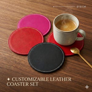 Op de afbeelding: Een set ronde leren onderzetters in roze, rood en zwart, elk voorzien van witte contraststiksels. De onderzetters hebben een diameter van 10 cm. Een keramische mok gevuld met koffie staat op een rode onderzetter, vergezeld door een gouden lepel.