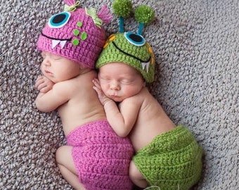 May include: Two babies wearing crocheted monster hats and matching diaper covers. The hats are green and pink with large eyes and teeth. The diaper covers are green and pink.