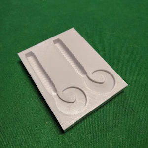 May include: A white, rectangular mold with two detailed cavities. Each cavity features a textured, elongated shape with a curved tail. The mold is likely for creating fishing lures or similar items.