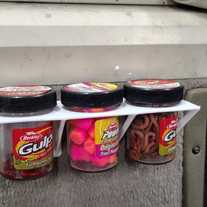 以下が含まれることがあります： 白いプラスチック製の棚に、黒い蓋のついた透明な瓶が3つ並んでいます。瓶には赤、ピンク、茶色の釣り餌が入っています。瓶には「Gulp」と「PowerBait」のブランド名が記載されたラベルが付いています。