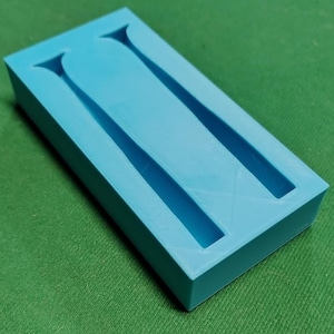 May include: A light blue rectangular mold with two long, narrow, parallel cavities. The mold has a smooth surface and is set against a green background. The cavities have a flared top.