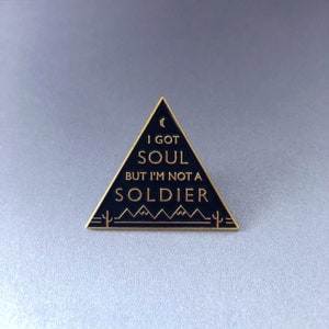 May include: A black triangle-shaped enamel pin with gold lettering. The pin reads "I got soul but I'm not a soldier" with a crescent moon and mountain range graphic.