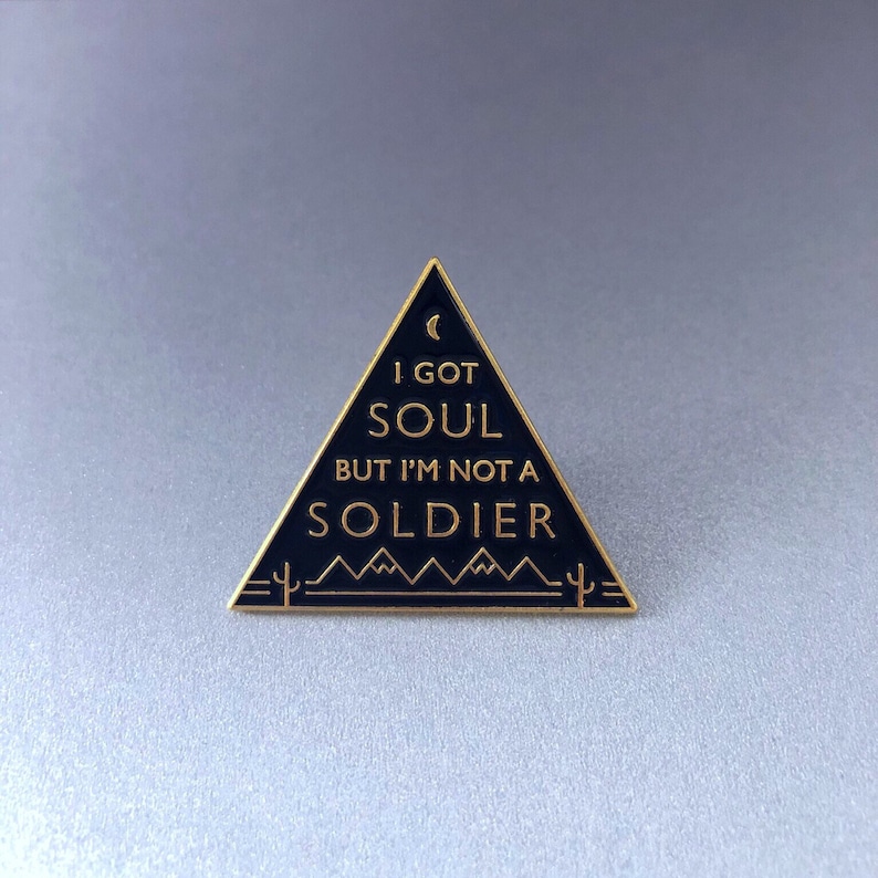 May include: A black triangle-shaped enamel pin with gold lettering. The pin reads "I got soul but I'm not a soldier" with a crescent moon and mountain range graphic.