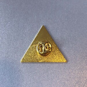 May include: A gold triangle-shaped pin with a pin back. The pin is blank.