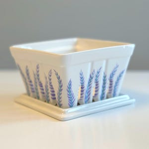 May include: A square, white ceramic planter with a matching square saucer. The planter is decorated with a repeating pattern of lavender-colored sprigs. The planter is designed for indoor or outdoor use.