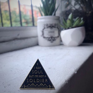 May include: A black and gold enamel pin shaped like a triangle with the text "I got soul but I'm not a soldier" inside. The pin has a crescent moon and cactus designs.