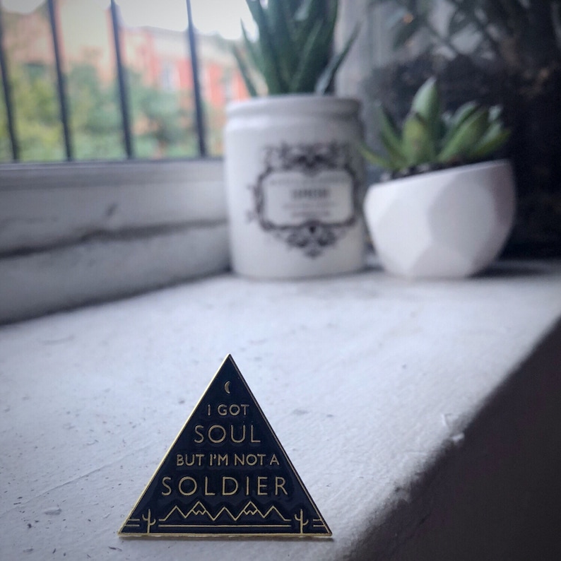 May include: A black and gold enamel pin shaped like a triangle with the text "I got soul but I'm not a soldier" inside. The pin has a crescent moon and cactus designs.
