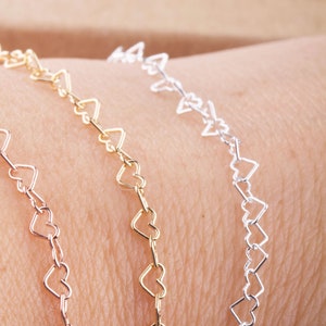 Puede incluir: Tres delicados brazaletes de cadena en forma de corazón en oro, plata y oro rosa. Los brazaletes están superpuestos en la muñeca de una persona.