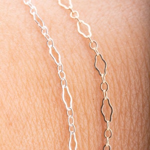 May include: Close-up of two delicate chain necklaces. One is silver-colored, the other is gold-colored. Both feature a repeating pattern of elongated diamond-shaped links. The chains are displayed against a skin-toned background.