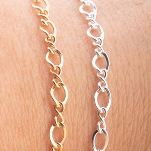 Könnte beinhalten: Nahaufnahme von zwei zarten Kettenarmbändern. Eines ist goldfarben, das andere silberfarben. Die Ketten haben ein ovales Gliederdesign und wirken schlicht und elegant. Die Armbänder werden vor einem hautfarbenen Hintergrund präsentiert.