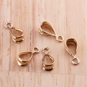 May include: Five gold-colored pendant bails, teardrop-shaped with a loop at the top for attaching to a chain or cord. The bails have a ribbed design on the lower portion and are displayed on a light wood surface.