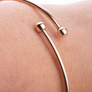 May include: A gold-colored open cuff bracelet with a minimalist design. The bracelet features two small, round ball accents at the open ends. The jewelry is shown on a person's arm, highlighting the sleek, modern style.