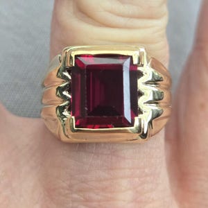 May include: A gold ring with a large, rectangular, deep red gemstone. The ring has a thick band with a textured design on the sides, and a gold bezel setting around the gemstone.