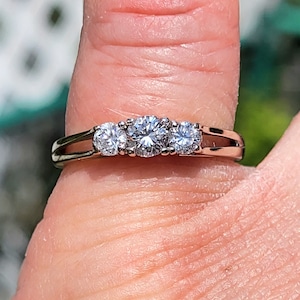 Puede incluir: Un anillo de compromiso de oro con tres piedras preciosas claras. El anillo tiene un diseño clásico, con tres piedras redondas engastadas juntas. El anillo se muestra en un dedo, con un fondo borroso.