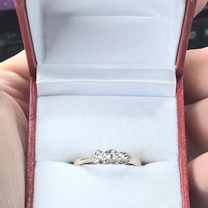 Puede incluir: Un anillo de oro con tres diamantes redondos engastados en fila. El anillo está en una caja de anillo de terciopelo rojo.