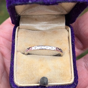 Pode incluir: Um anel de banda de prata com pequenos diamantes redondos engastados em uma fila ao redor da banda. O anel está em uma caixa de anel de veludo roxo.