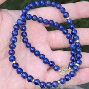 May include: A beaded necklace featuring deep blue lapis lazuli stones. The necklace has gold-colored spacers and a gold clasp. The round beads are strung together to create a classic jewelry piece.