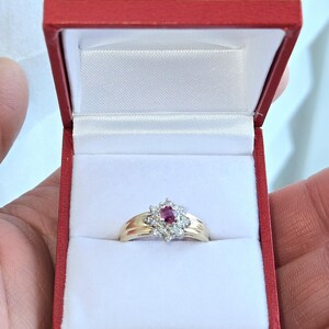 Op de afbeelding: Een gouden ring met een centrale robijn omringd door kleinere diamanten, gepresenteerd in een rode sieradendoos met een witte satijnen binnenkant. Het bloemendesign en de glans van de ring worden benadrukt.