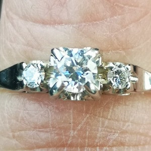 Puede incluir: Un anillo de plata con tres diamantes redondos. El diamante central es más grande que los dos diamantes laterales.