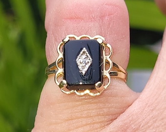 May include: A gold ring featuring a black square stone set within an ornate frame. A small diamond is set in the centre of the black stone.