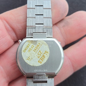 May include: A vintage silver-toned wristwatch with a rectangular face and a linked metal band. The watch has a sticker on the back with text that reads "SERIES 25" and "USE CELL 565". The watch is held in a hand.