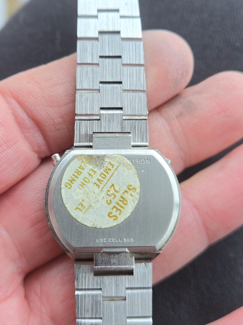 May include: A vintage silver-toned wristwatch with a rectangular face and a linked metal band. The watch has a sticker on the back with text that reads "SERIES 25" and "USE CELL 565". The watch is held in a hand.
