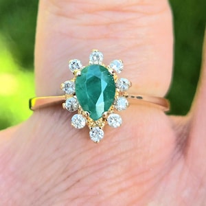 May include: A gold ring with a pear-shaped emerald gemstone surrounded by a halo of smaller diamonds.