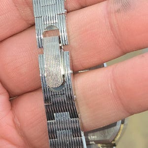 May include: Close-up of a silver-toned watch band with a textured, woven design. The band is connected with a clasp. The watch band is held by a person's hand.
