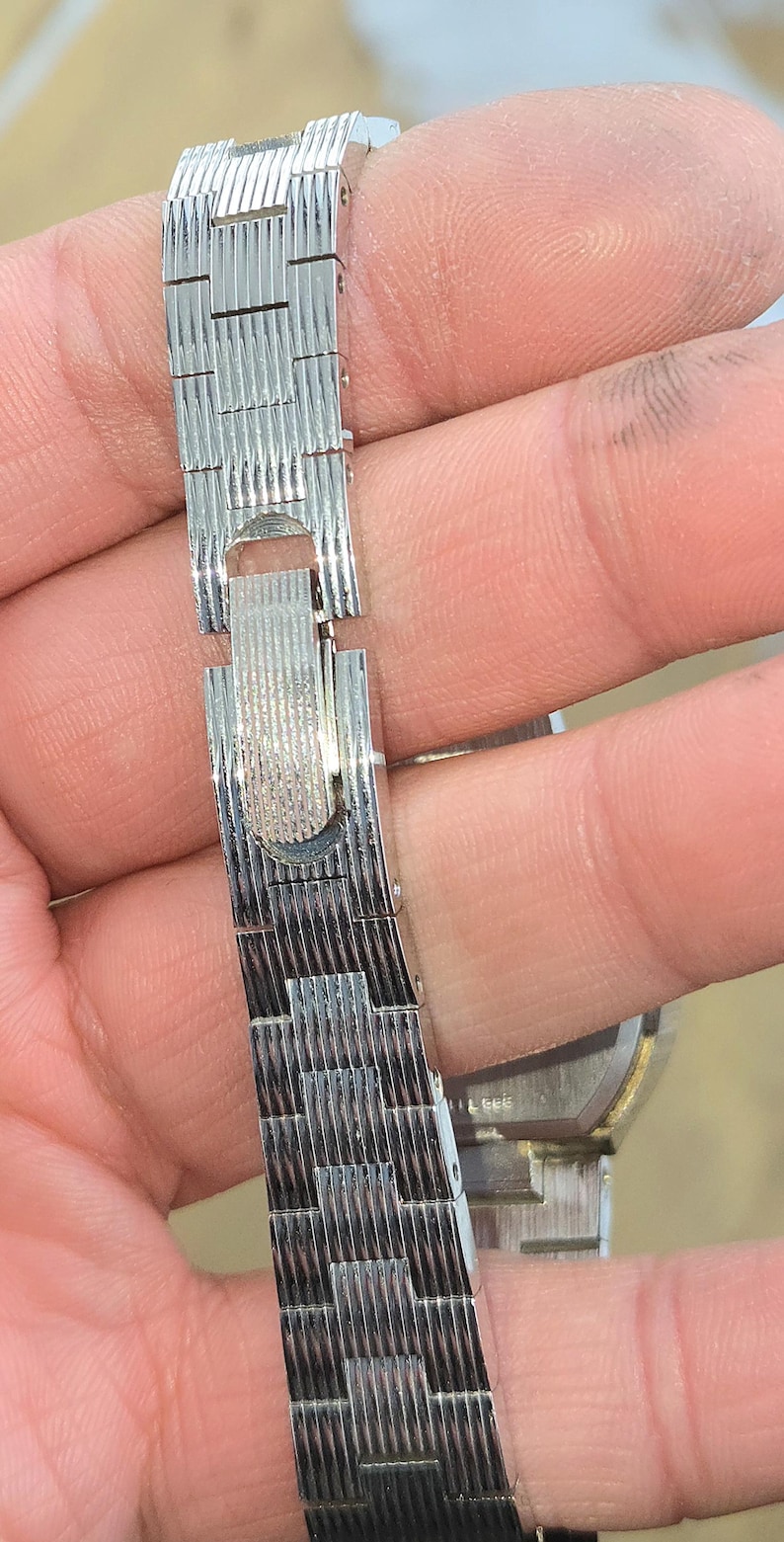 May include: Close-up of a silver-toned watch band with a textured, woven design. The band is connected with a clasp. The watch band is held by a person's hand.