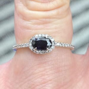 May include: A silver ring featuring a dark oval gemstone surrounded by a halo of smaller, clear stones. The band is also adorned with small, clear stones. The ring is set against a blurred background.