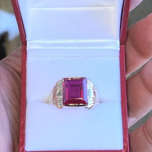 Peut inclure: Une bague en argent avec une grosse pierre précieuse rectangulaire de couleur rubis. La bague est présentée dans un écrin rouge avec un intérieur blanc. Le design de la bague comprend des éléments décoratifs sur les côtés.