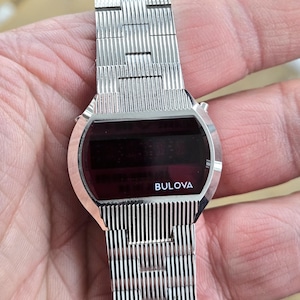 May include: A silver-toned Bulova digital watch with a rectangular red display. The watch features a textured metal band and the Bulova logo is visible on the watch face. The watch is a vintage style.