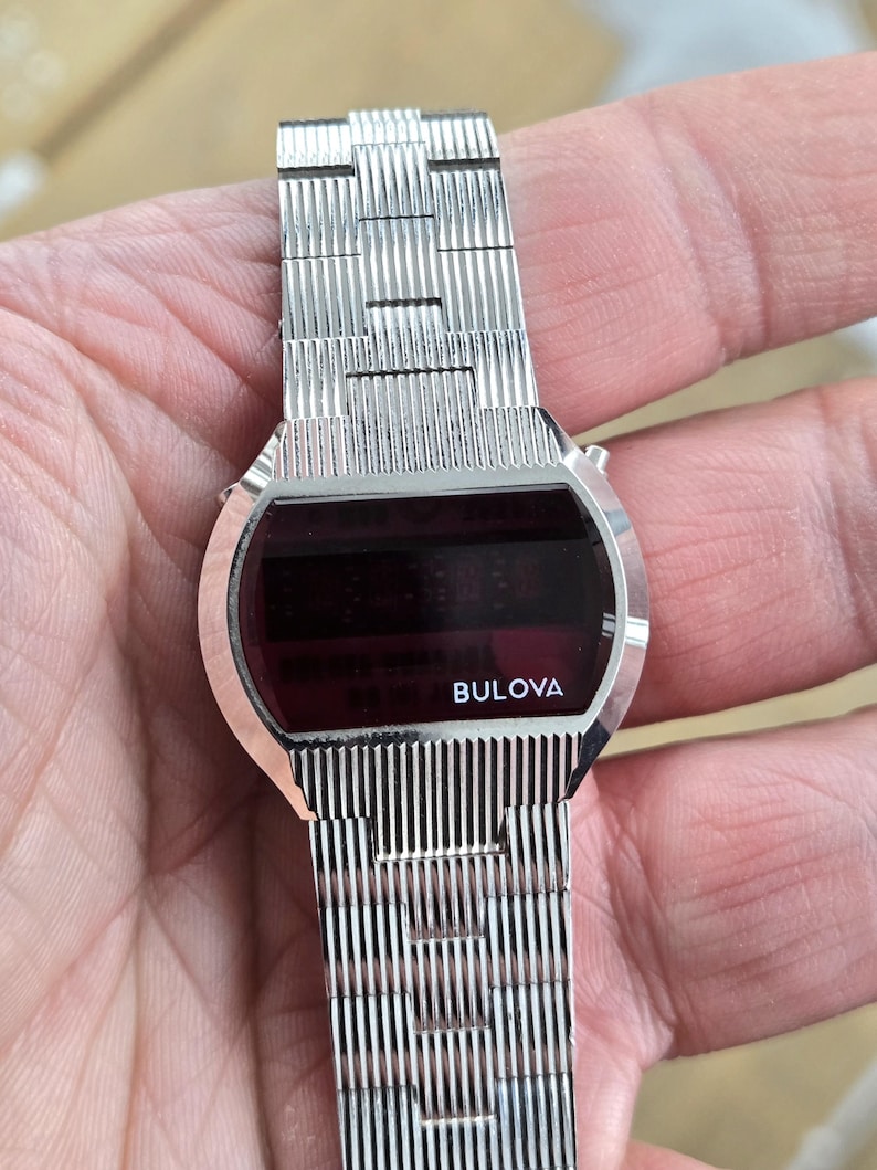 May include: A silver-toned Bulova digital watch with a rectangular red display. The watch features a textured metal band and the Bulova logo is visible on the watch face. The watch is a vintage style.