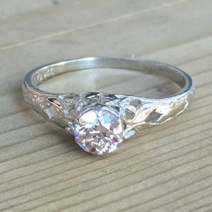May include: A silver engagement ring with a large, round, clear gemstone. The band features intricate floral filigree and the inscription "14K". The ring is set against a light wood grain background.