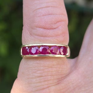 May include: A gold ring set with five oval-cut, deep red rubies. The gemstones are arranged in a line, forming a continuous band. The ring is displayed on a finger, showcasing the jewellery's design and craftsmanship.