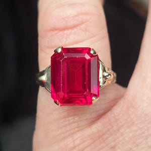 Könnte beinhalten: Ein goldener Ring mit einem großen, rechteckigen, facettierten, tiefroten Edelstein. Der Ring hat ein verziertes Band, und der Edelstein ist mit vier Krappen gefasst. Der Ring wird an einem Finger gezeigt.