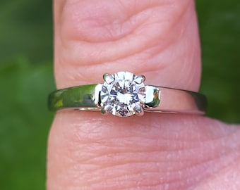 Könnte beinhalten: Ein silberner Ring mit einem großen, runden, klaren Edelstein. Der Ring befindet sich an einem Finger vor einem verschwommenen grünen Hintergrund. Der Edelstein ist der Mittelpunkt und reflektiert das Licht.
