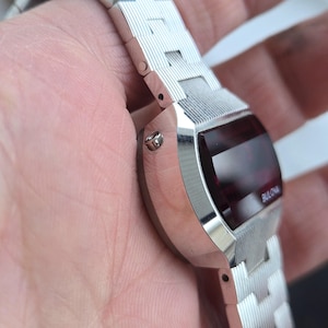 May include: A silver-toned Bulova digital watch with a rectangular red display. The watch has a matching silver band with a textured design. The word "Bulova" is visible on the watch face.