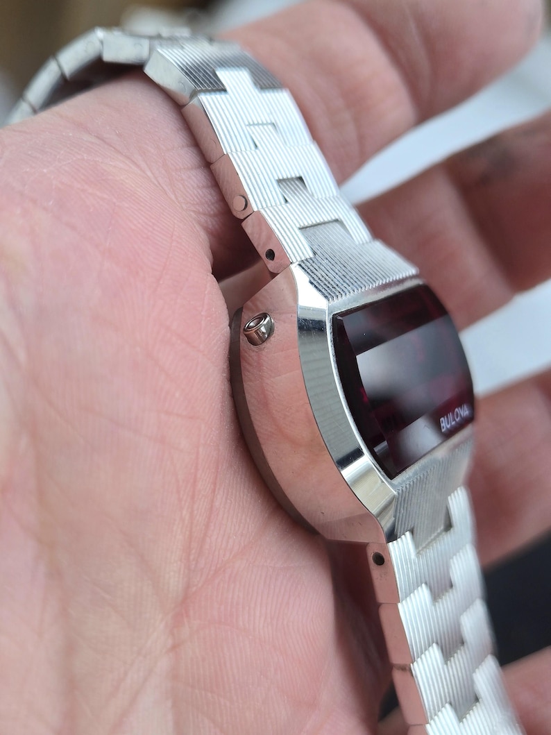 May include: A silver-toned Bulova digital watch with a rectangular red display. The watch has a matching silver band with a textured design. The word "Bulova" is visible on the watch face.