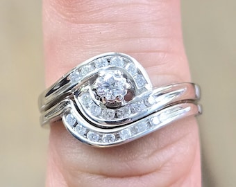 May include: A silver engagement ring and wedding band set. The engagement ring has a central, round-cut diamond surrounded by smaller stones. The band has a wave design with small diamonds. The rings are on a finger.