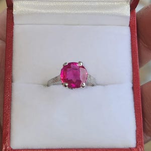 Puede incluir: Un anillo de plata con una gran piedra preciosa rosa brillante de talla ovalada. El anillo está engastado en un engaste de cuatro puntas, presentado en una caja de joyería roja y blanca. La banda presenta detalles delicados.