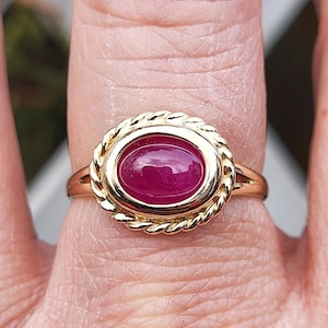 May include: A gold ring with a red oval gemstone set in a rope-like border.