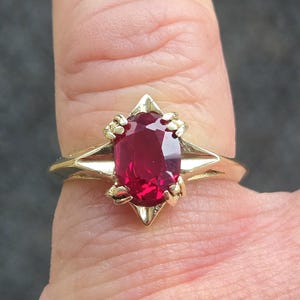 May include: A gold ring featuring a large, oval-cut ruby gemstone. The ruby is a vibrant red color and is set in a gold band with a unique, geometric design. The ring is a piece of jewelry.